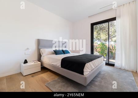 Das moderne, stilvolle Schlafzimmer in einer luxuriösen mediterranen Villa verfügt über ein minimalistisches Bett, weiße Möbel, luftige Vorhänge und raumhohe Fenster Stockfoto