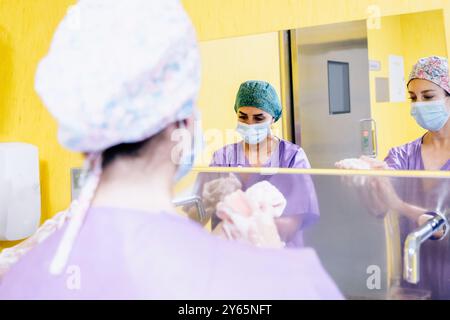 Zwei Chirurgen in Peelings und Masken bereiten sich auf die Operation vor und konzentrieren sich auf ihre Arbeit hinter einem Schutzglas in einem hell beleuchteten Krankenhausraum Stockfoto