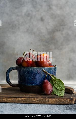 Bio-reife Pflaumen in einer rustikalen blauen Keramikschale, ergänzt durch ein frisches Blatt, vor einem strukturierten grauen Hintergrund. Perfekt zur Illustration von Farm-t Stockfoto