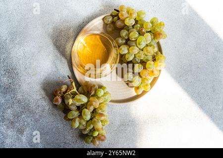 Blick von oben auf ein erfrischendes Glas Weißwein neben Gruppen frischer grüner Trauben, präsentiert auf einem Holzteller auf einem strukturierten grauen Hintergrund, il Stockfoto