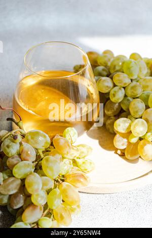In einer hellen, sonnendurchfluteten Umgebung paart sich ein Glas Weißwein mit frischen Trauben und wirft einen weichen Schatten auf eine strukturierte Oberfläche. Diese einladende sce Stockfoto