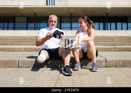 Ein Glatzkopf mit amputiertem Bein stellt seine Prothese mit Hilfe seiner Tochter ein, beide in Sportkleidung gekleidet, und sitzt auf einer Treppe vor einem modernen Stockfoto