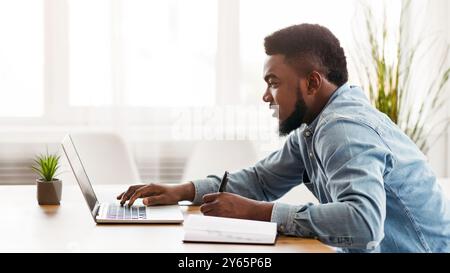 Schwarzer Millennial-Angestellter, der im Büro einen Laptop benutzt und Notizen macht Stockfoto