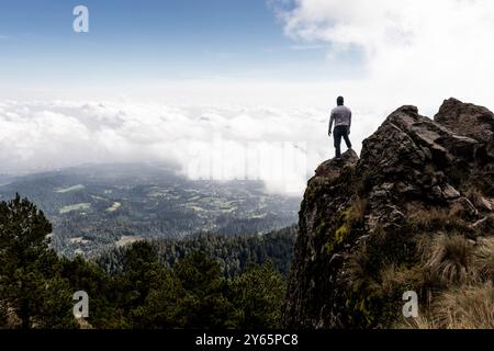 Ein einsamer Wanderer steht auf einem zerklüfteten Berggipfel und blickt über ein Tal, das von einer dicken Wolkenschicht umgeben ist, und zeigt die Majestät und Einsamkeit der Natur Stockfoto