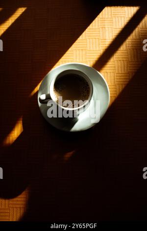 Das Morgenlicht wirft geometrische Schatten auf einen Holztisch, der eine Tasse dampfenden Kaffee hält, und ruft eine ruhige, besinnliche Stimmung hervor. Keine Leute im fram Stockfoto