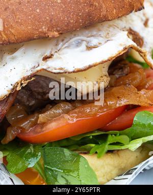 Nahaufnahme eines köstlichen Gourmet-Burgers mit saftigem Patty, geschmolzenem Käse, Speck, frischen Tomatenscheiben, Rucola, und ein Spiegelei, alles in Sandwichform Stockfoto