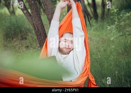 Ein zufriedener junger Mann dehnt sich entspannt in einer leuchtend orangen Hängematte zwischen Bäumen aus, ein Laptop an seiner Seite und verbindet Arbeit und Spiel in einer ruhigen Atmosphäre Stockfoto