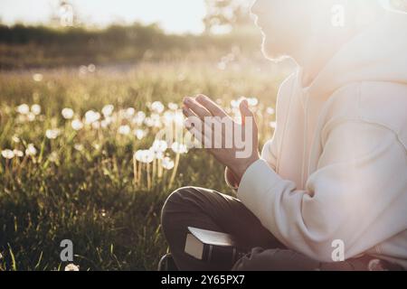 Seitenansicht eines nicht erkennbaren Mannes, der draußen sitzt, die Bibel liest und in einer natürlichen, sonnendurchfluteten Umgebung betet. Die Szene zeigt eine friedliche Spiritua Stockfoto