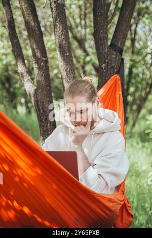 Ein junger Mann entspannt sich in einer leuchtend orangen Hängematte im Freien, konzentriert auf seinen Laptop, umgeben von der ruhigen Atmosphäre eines üppigen Waldes Stockfoto