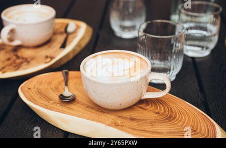 Blick von oben auf zwei Keramikbecher Kaffee mit Latte Art, auf einzigartigen Holztabletts neben klaren Wassergläsern, dargestellt in einem gemütlichen Café-Ambiente. Stockfoto