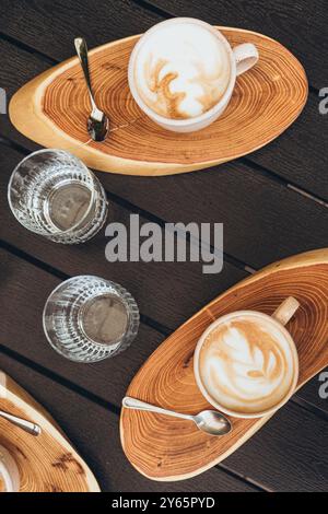 Blick von oben auf zwei Cappuccinos mit kunstvollen Schaummustern, serviert auf Holztellern neben klaren Gläsern Wasser, auf einem dunklen Holztisch. Stockfoto