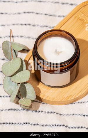 Eine ruhige Einrichtung mit einer aromatischen weißen Kerze in einem kleinen braunen Glasgefäß, begleitet von frischen Eukalyptusblättern auf gestreiftem Leinen Stockfoto