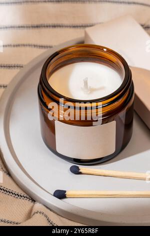 Nahaufnahme einer unbeleuchteten Duftkerze in einem stilvollen braunen Glasgefäß, ergänzt durch Holzhölzer, vor einem gestreiften Stoffhintergrund Stockfoto