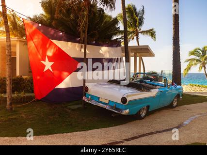 Eine malerische Szene aus Varadero, Kuba, mit einem blauen Oldtimer neben einer großen kubanischen Flagge, mit Palmen und dem Meer im Hintergrund Stockfoto