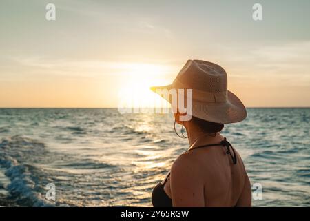 Rückansicht einer nicht erkennbaren Frau, die den Sonnenuntergang an einem Varadero-Strand in Kuba beobachtet, mit einem Strohhut und einem ruhigen Meer im Hintergrund. Stockfoto