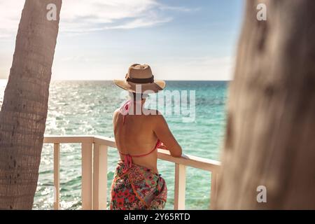 Rückansicht einer nicht erkennbaren Frau mit Strohhut und rotem Bikinioberteil, die das Karibische Meer von einem Balkon in Varadero, Kuba, bewundert. Stockfoto