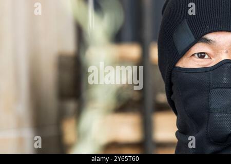 Nahaufnahme eines mysteriösen asiatischen Mannes in schwarzer Sturmhaube, mit einem Auge sichtbar, das Geheimhaltung oder Schutz symbolisiert. Dahinter deutet der verschwommene Hintergrund auf t an Stockfoto