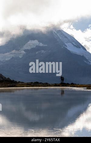 Rückblick auf die nicht erkennbare Silhouette eines Paares, das an einem ruhigen See steht, mit schneebedeckten Bergen, die sich im Wasser spiegeln Stockfoto