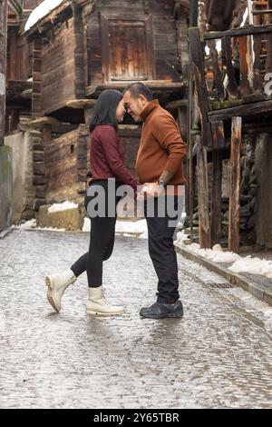 Seitenansicht eines asiatischen Paares, das liebevoll die Stirn berührt, während Weihnachten auf einer kopfsteingepflasterten Straße in Zermatt steht, umgeben von altem Holz Stockfoto