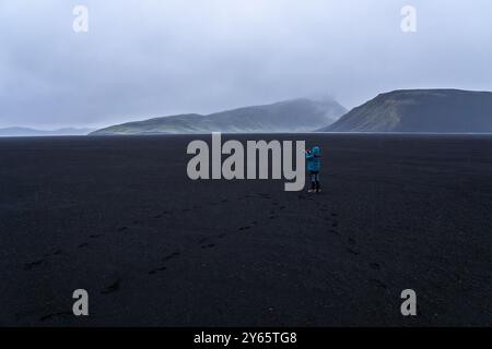 Ein Alleinreisender steht auf dem riesigen schwarzen vulkanischen Sand des isländischen Hochlands und fängt die unheimliche und nebelige Landschaft unter einem bewölkten Himmel ein Stockfoto