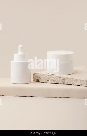 Dieses Bild zeigt moderne Hautpflegeprodukte wie einen Serumtropfer und einen Cremebecher, die auf einer einfachen Natursteinplatte gegen eine weiche Be angeordnet sind Stockfoto