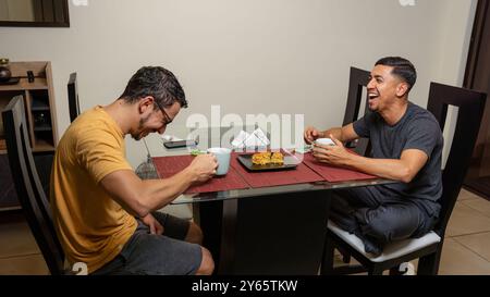 Ein fröhliches schwules Paar teilt ein angenehmes Essen zusammen und führt in gemütlicher Atmosphäre zu einer entspannten Unterhaltung und Lachen, wobei LGBTQI hervorgehoben wird Stockfoto