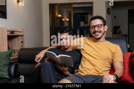Ein liebevolles schwules Paar teilt einen angenehmen Moment auf der Couch, wobei ein Mann ein Buch liest und der andere ihn freudig umhüllt Stockfoto
