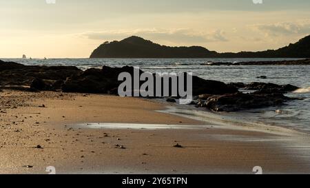 Ruhiger Blick auf den Sonnenuntergang an den Sandstränden des Golfs von Papagayo in Costa Rica, mit goldenem Licht, felsigen Formationen und einem weit entfernten Segelboot. Stockfoto