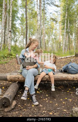 Eine Mutter mit zwei Töchtern, die auf einem Baumstamm im Wald sitzen und einen Moment der Freude während eines Wanderabenteuers teilen, umgeben von einem Birkenwald Stockfoto