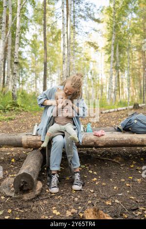 Eine Mutter und ihre kleine Tochter genießen eine Pause während einer Wanderung in einem üppigen Wald. Die Mutter küsst ihr Kind zärtlich, das auf einem Baumstamm sitzt, surr Stockfoto