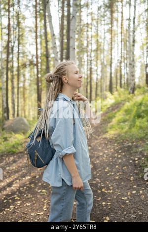 Eine junge Frau mit langen Dreadlocks und einem Rucksack blickt nachdenklich auf, während sie auf einem Waldweg spaziert und vermittelt ein Gefühl von Abenteuer und Ruhe Stockfoto