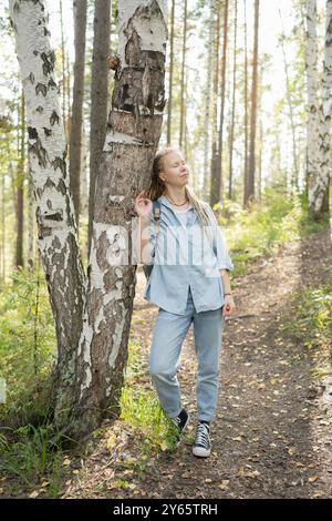 Eine junge Frau mit langen Dreadlocks und einem Rucksack blickt nachdenklich auf, während sie auf einem Waldweg spaziert und vermittelt ein Gefühl von Abenteuer und Ruhe Stockfoto