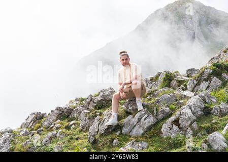 Ein junger Mann sitzt nachdenklich auf einem felsigen Hang in Asturien, umgeben von nebeliger Bergluft, und blickt nachdenklich von der Kamera weg. Stockfoto