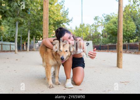 Eine lächelnde junge Frau umarmt liebevoll einen Shetland-Schäferhund, während sie ein Selfie in einem sonnigen Park macht, das eine Verbindung zwischen Haustier und Besitzer zeigt Stockfoto