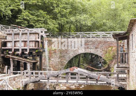 Eine alte Holzwassermühle befindet sich neben einer Ziegelbogenbrücke, umgeben von lebendigen grünen Wasserkaskaden aus dem Mühlenrad, die Energie c darstellen Stockfoto