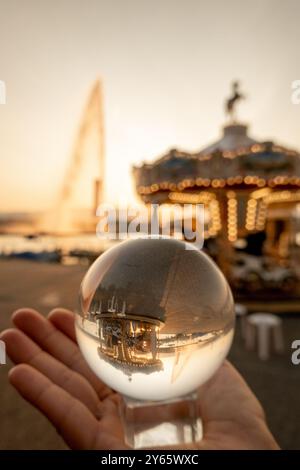 Eine abgeschnittene, nicht erkennbare Person hält eine Kristallkugel, die den Genfer Jet d'Eau reflektiert, und ein Karussell bei Sonnenuntergang, mit lebendigen Reflexen und Stockfoto