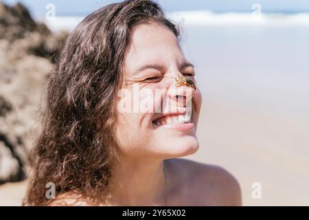 Eine glückliche junge Frau, die einen sonnigen Strandtag genießt, mit Krabben auf der Nase lacht und unbeschwerten Sommerspaß verkörpert Stockfoto