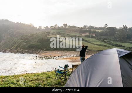 Rückansicht eines nicht erkennbaren männlichen Touristen, der neben einem Zelt steht und eine Küstenlandschaft genießt, mit Campingausrüstung im Vordergrund. Stockfoto