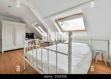 Helles und luftiges Schlafzimmer im Dachgeschoss mit weißem Eisenrahmen, Dachfenster, Holzboden und einem einfachen Hocker. Stockfoto