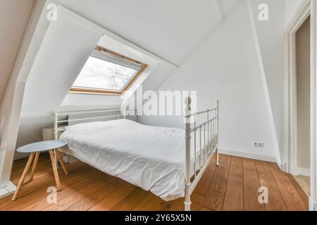 Ein minimalistisches Schlafzimmer im Dachgeschoss mit weißem Eisenbett, einem kleinen blauen Beistelltisch und einem Dachfenster mit Tageslicht und Blick auf den Himmel. Stockfoto