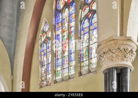 Eine Reihe von wunderschönen, mehrfarbigen Buntglasfenstern lässt Licht in eine alte Kirche und zeigt religiöse Bilder neben einer kunstvollen Säule. Stockfoto