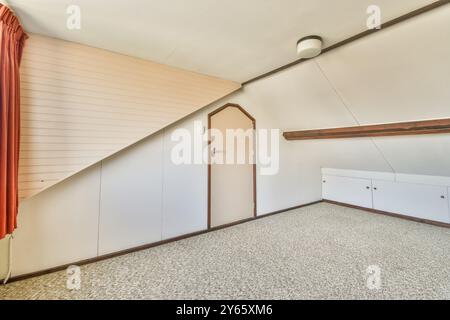 Ein luftiger und heller Dachboden mit schräger Decke, eingebauten Abstellschränken und flauschigem Teppichboden mit rotem Vorhang. Stockfoto