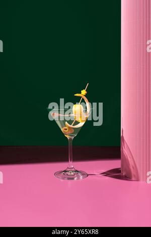 Ein klassischer, trockener Martini-Cocktail, elegant präsentiert mit Oliven und Zitronenschalen vor einem kontrastierenden grünen und rosa Hintergrund. Stockfoto