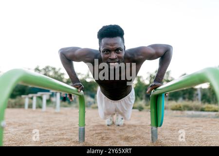 Ein fokussierter, muskulöser Mann nimmt an einem Calisthenics-Workout Teil und demonstriert einen Push-up auf parallelen Bars in einem sonnendurchfluteten Outdoor-Park, der Stärke und Stärke hervorhebt Stockfoto