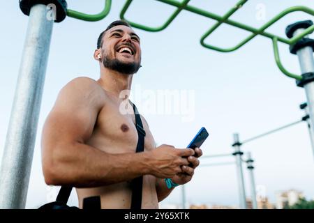 Ein fröhlicher Mann mit Bart greift auf sein Smartphone und ruht sich in einem Calisthenics-Trainingspark aus. Seine Pause während eines Workouts unterstreicht die perfekte Mischung Stockfoto