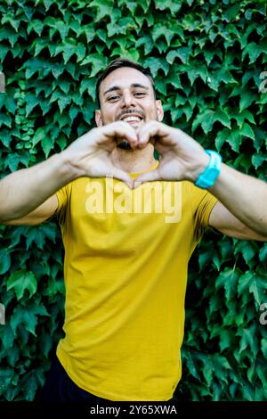 Ein lächelnder junger Mann in einem gelben Hemd bildet mit seinen Händen vor einem üppigen, grünen Efeu-Hintergrund eine Herzform und zeigt eine verspielte und liebevolle Geste Stockfoto