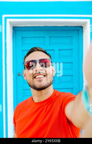 Fröhlicher junger Mann mit Bart, der draußen vor einer hellblauen Tür ein Selfie macht, Sonnenbrille und ein rotes T-Shirt trägt Stockfoto
