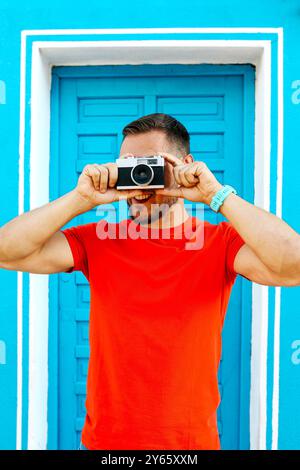Ein junger Mann in einem knallroten Hemd nimmt mit einer alten Kamera ein Foto auf und steht vor einer leuchtend blauen Wand mit türkisfarbenem Türrahmen Stockfoto