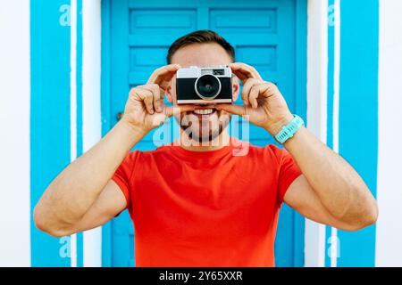 Ein fröhlicher Mann in einem knallroten Hemd macht spielerisch ein Foto mit einer Vintage-Kamera vor einer markanten blauen Tür Stockfoto
