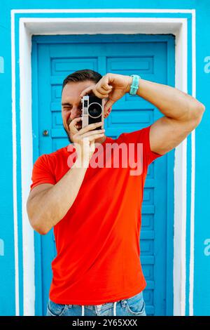 Der fröhliche junge Mann mit rotem T-Shirt und blauer Jeans macht gerne Fotos mit einer alten Kamera vor einer leuchtenden blauen Tür Stockfoto